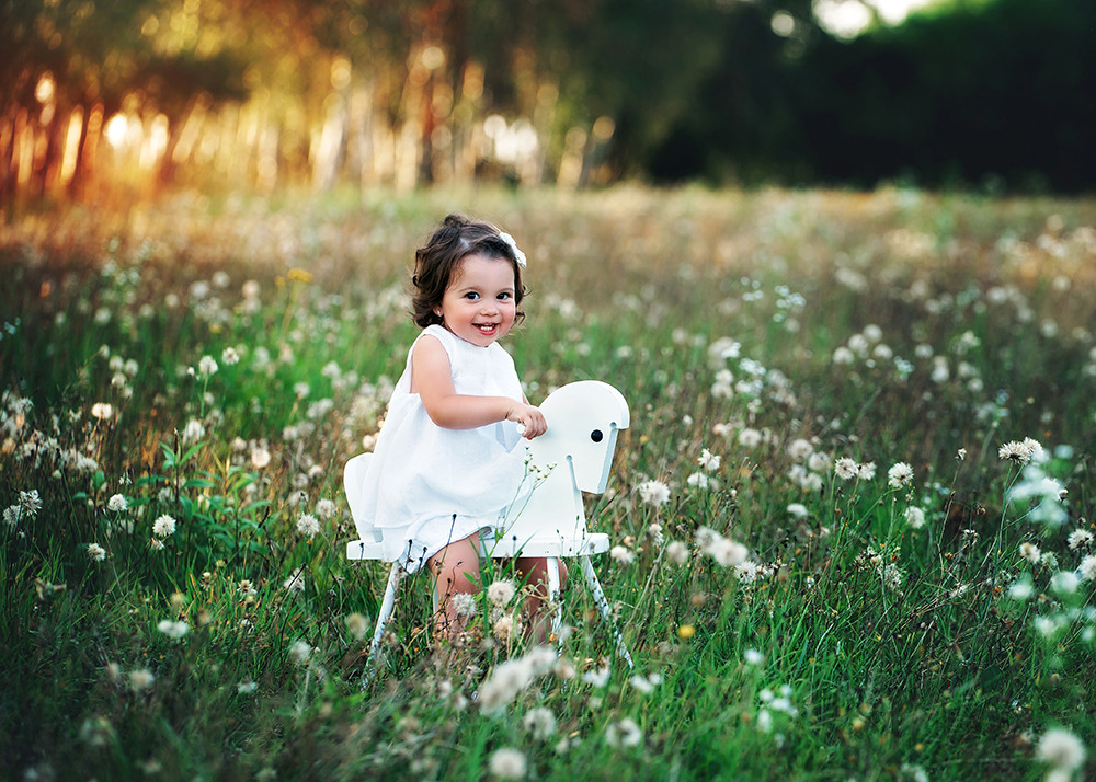Uśmiechnięta dziewczynka w białej sukience siedzi na białym, drewnianym koniku na biegunach, otoczona kwitnącą łąką w ciepłym, popołudniowym świetle.