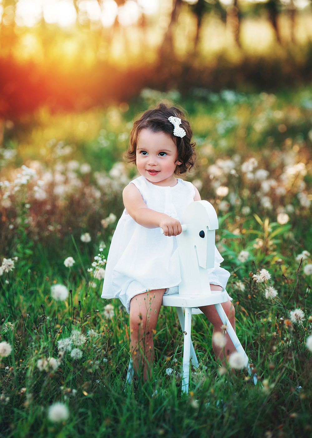 Dziewczynka w białej sukience i z kokardą we włosach siedzi na białym koniku na biegunach na łące z dmuchawcami, w tle rozmyte drzewa i zachodzące słońce.