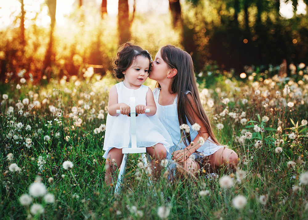 Dwie dziewczynki w białych sukienkach na łące z dmuchawcami, starsza całuje młodszą w policzek, młodsza siedzi na białym drewnianym koniku na biegunach.
