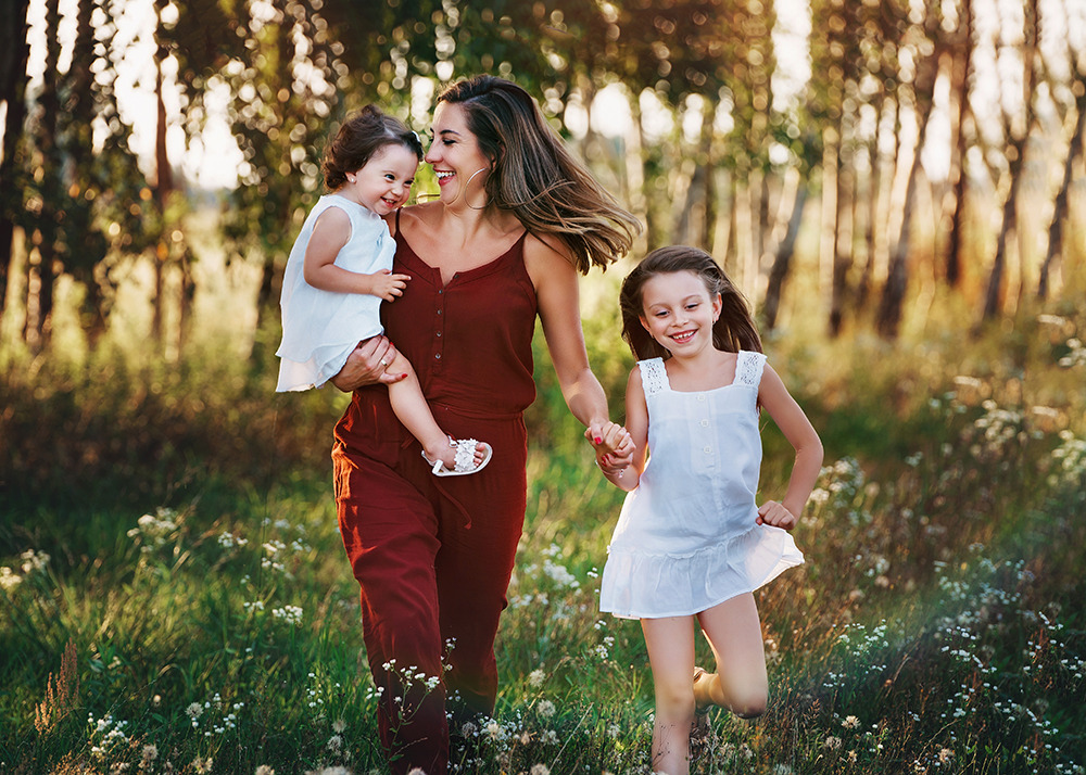 Uśmiechnięta mama w bordowym kombinezonie biegnie przez łąkę z dwiema córkami w białych sukienkach, trzymając jedną na rękach, a drugą za rękę, w tle drzewa i zachodzące słońce.