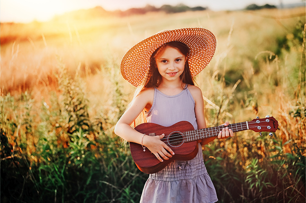 Dziewczynka w słomkowym kapeluszu gra na ukulele w trawie w promieniach zachodzącego słońca.