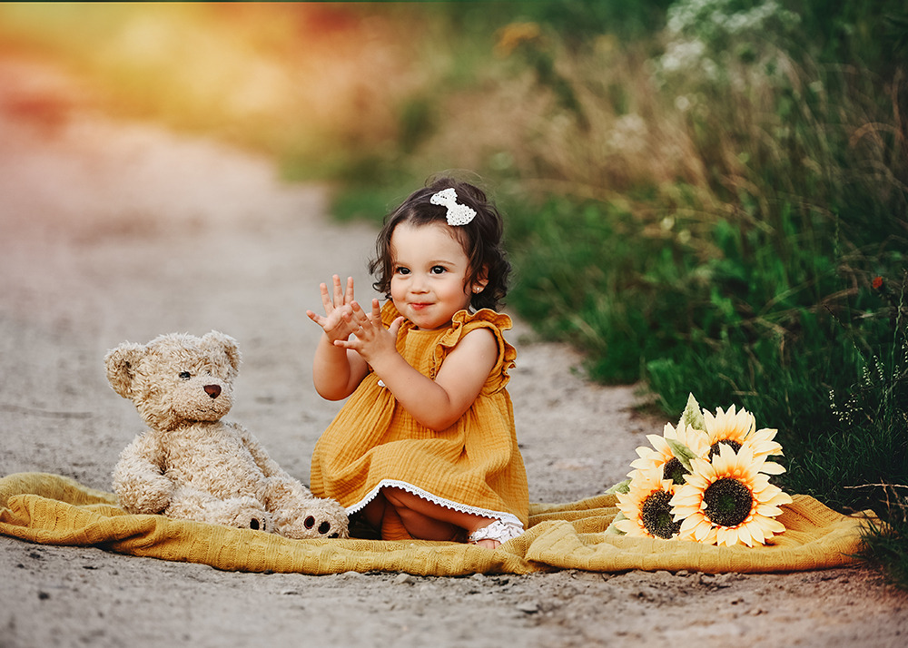 Uśmiechnięta dziewczynka w musztardowej sukience siedzi na żółtym kocu obok pluszowego misia i bukietu słoneczników, z podniesionymi rączkami na tle polnej ścieżki.