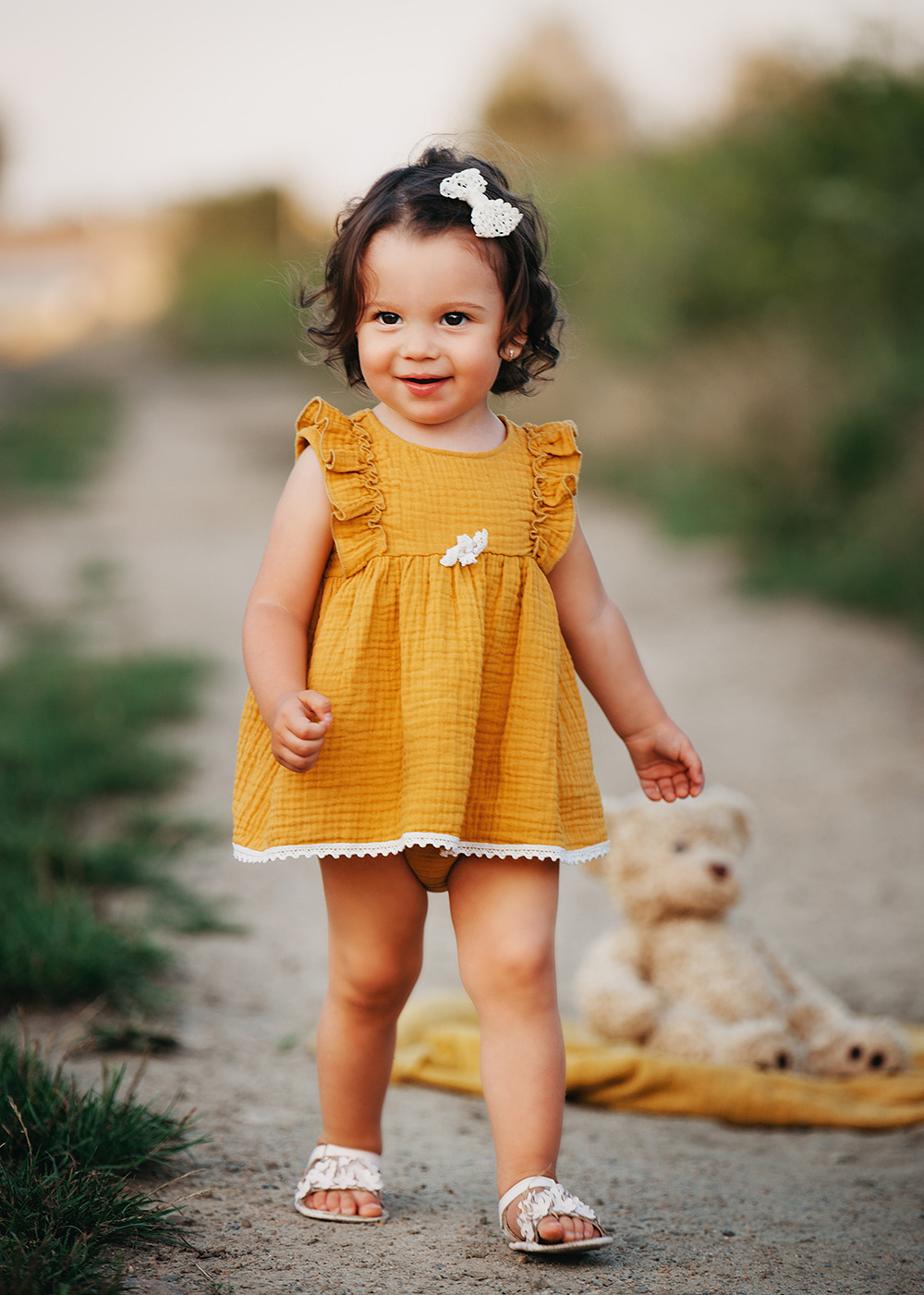Uśmiechnięta dziewczynka w musztardowej sukience z falbankami i kokardą we włosach, spacerująca po polnej drodze, z pluszowym misiem w tle.