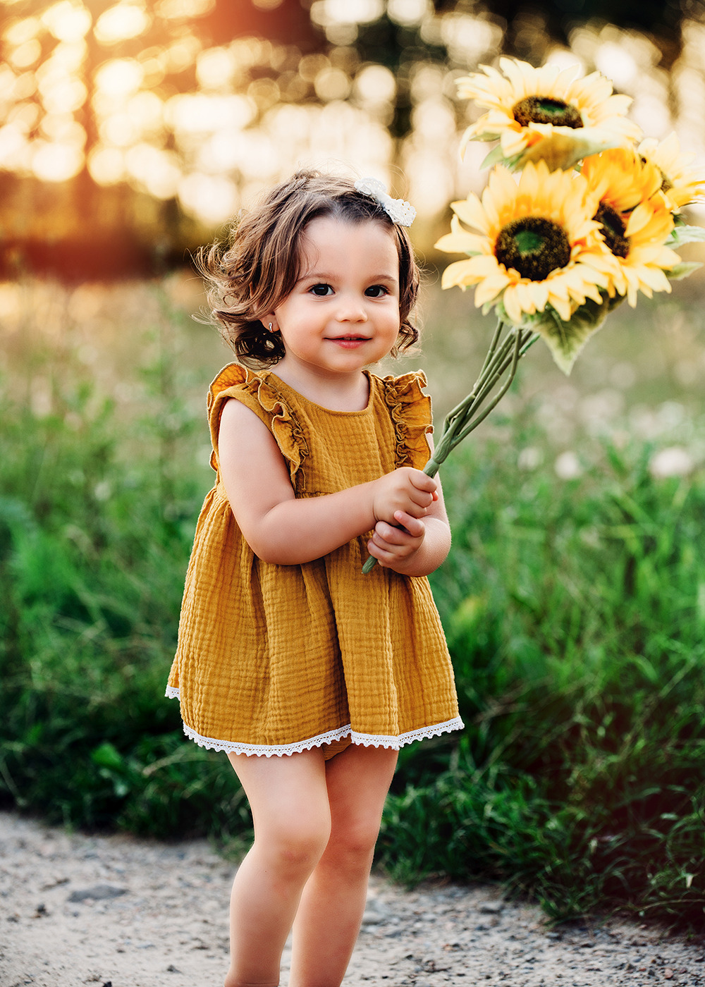 Uśmiechnięta dziewczynka w musztardowej sukience z falbankami trzyma bukiet słoneczników na tle zieleni i zachodzącego słońca.