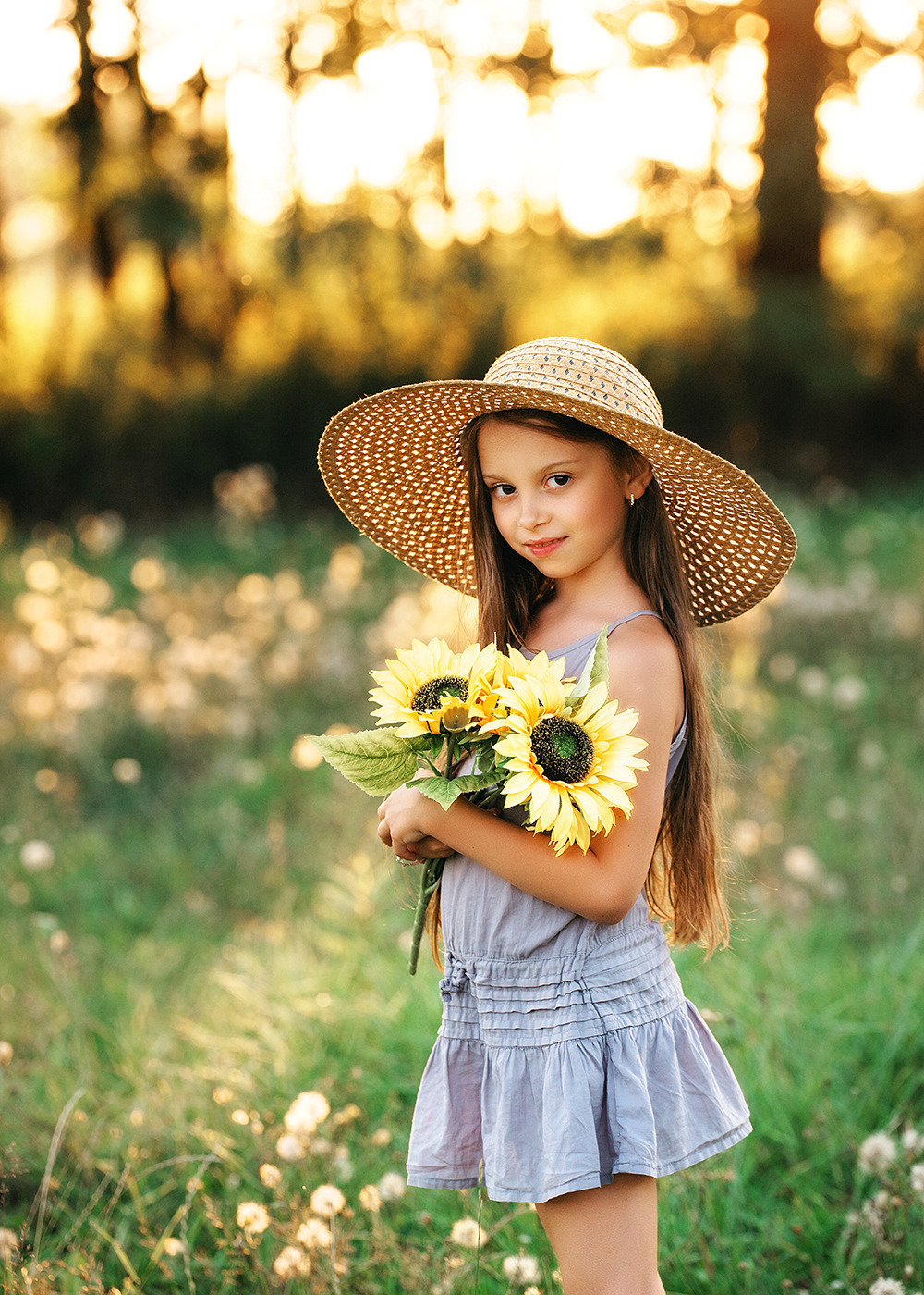 Uśmiechnięta dziewczynka w słomkowym kapeluszu trzyma bukiet słoneczników na łące w złotym świetle.