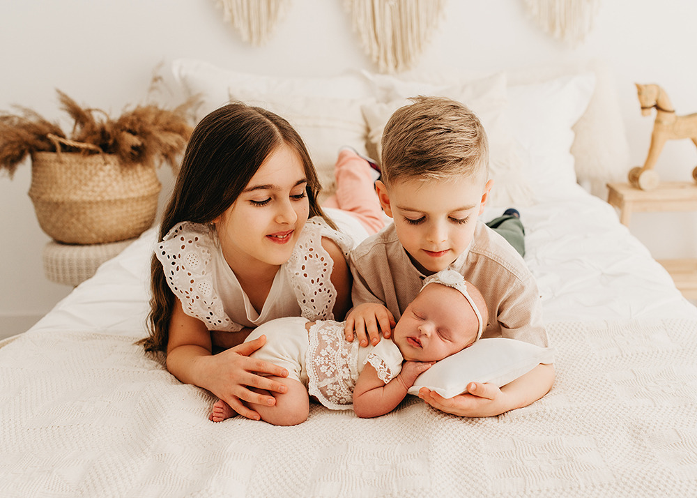 Rodzeństwo leżące na łóżku z noworodkiem, dziewczynka w białej bluzce z ażurowymi rękawami, chłopiec w beżowej koszuli, dziecko w kremowym ubranku z koronką, jasne wnętrze z dekoracjami boho.