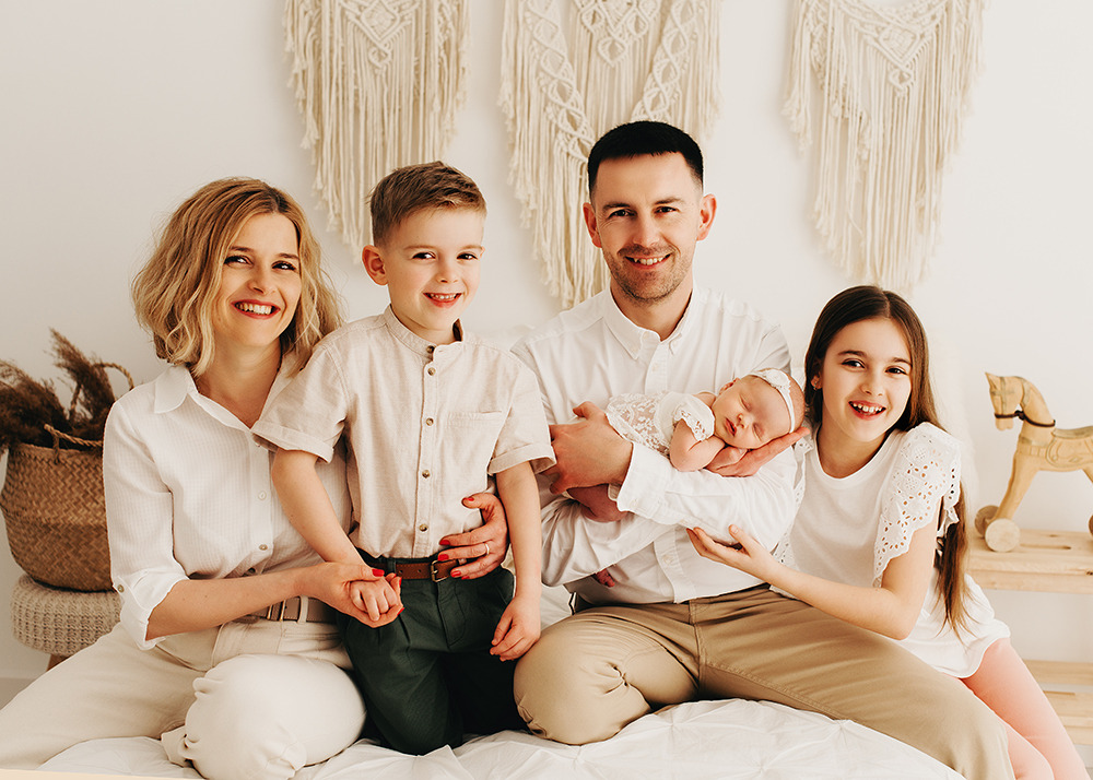 Uśmiechnięta rodzina z trójką dzieci, w tym noworodek w ramionach ojca, pozująca na białym tle z makramami i drewnianym koniem na biegunach.