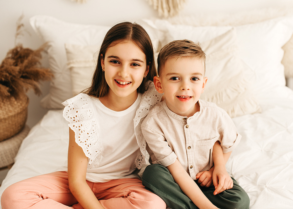 Uśmiechnięta dziewczynka w białej bluzce z koronkowymi rękawami siedzi obok uśmiechniętego chłopca w beżowej koszuli na białej pościeli, patrząc w obiektyw.