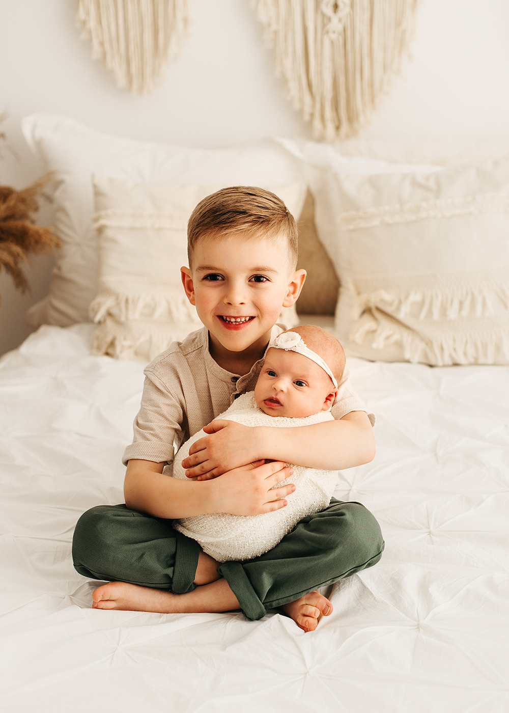 Uśmiechnięty chłopiec trzyma na kolanach zawinięte w biały kocyk niemowlę z białą opaską na głowie, siedząc na białej pościeli w pokoju z dekoracjami makramowymi na ścianie.