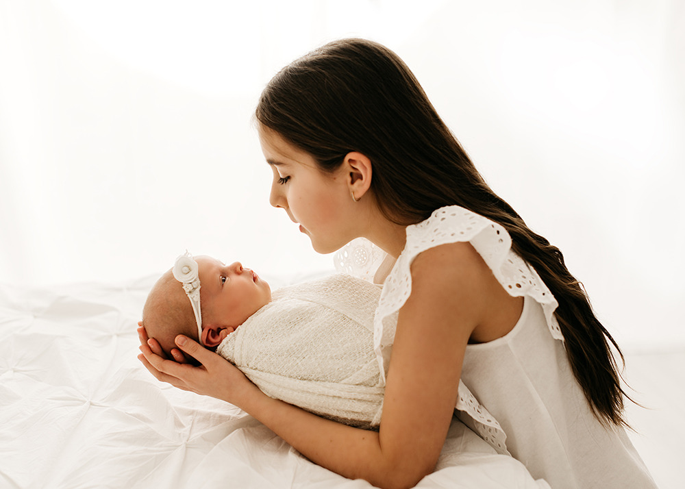 Młodsza siostra w białej bluzce z haftowanymi falbankami spogląda z czułością na zawinięte w kremowy kocyk niemowlę z opaską z kwiatkiem na głowie, leżące na białej pościeli.