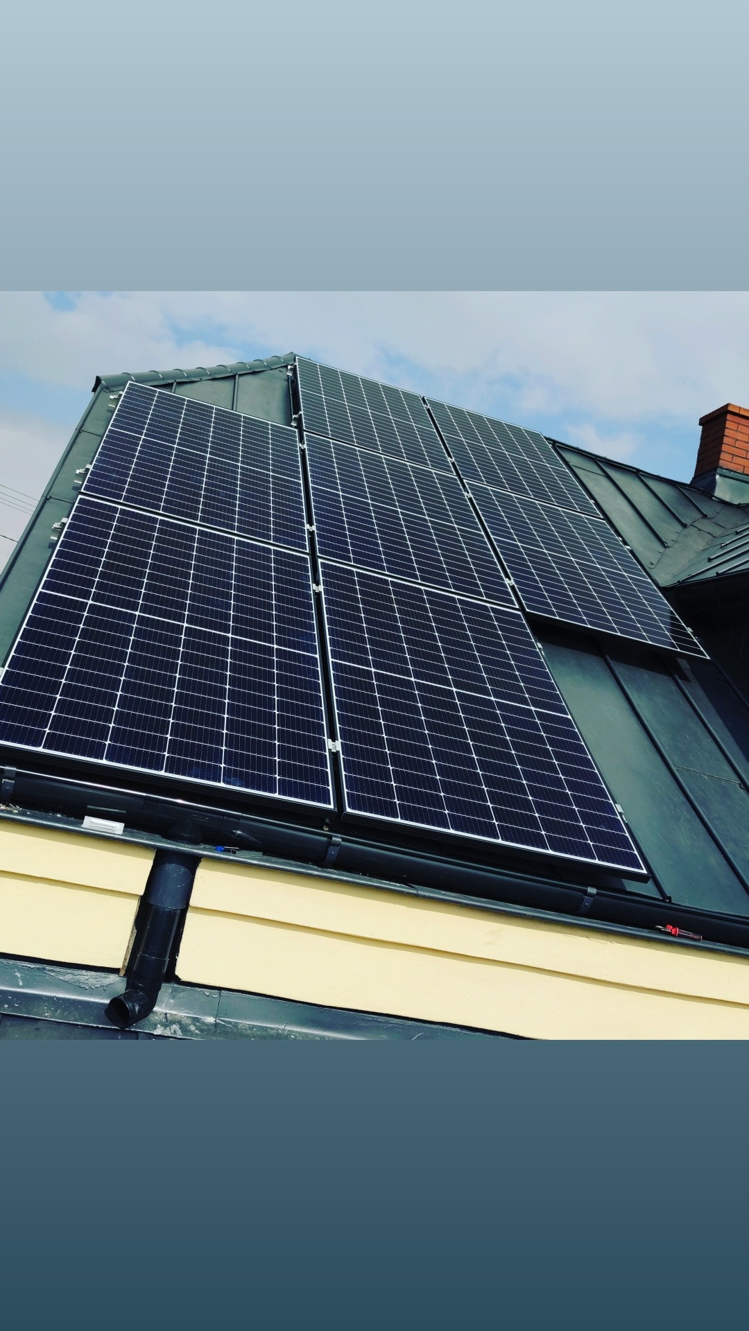 Panel słoneczny zamontowany na blaszanym dachu domu z widocznym kominem i rynną.