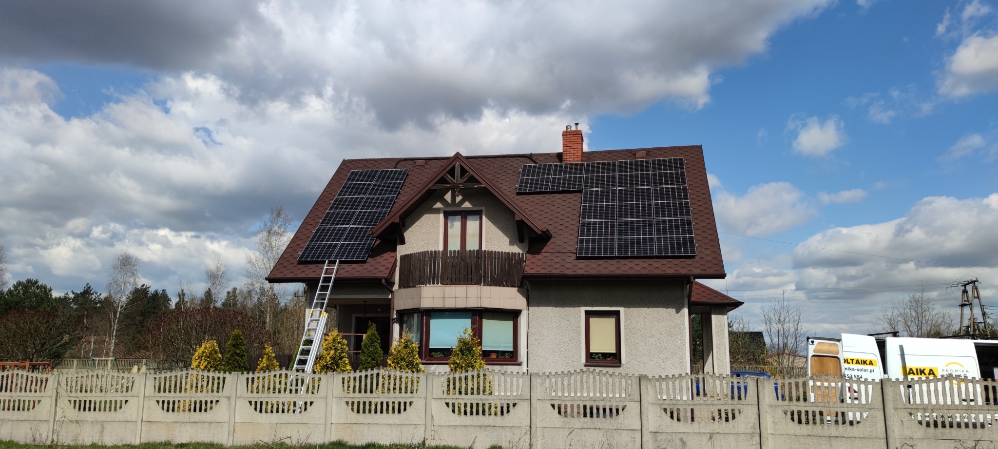 Dom jednorodzinny z panelami fotowoltaicznymi na dachu, drabina oparta o dach, widoczne częściowo auto dostawcze firmy instalującej panele.