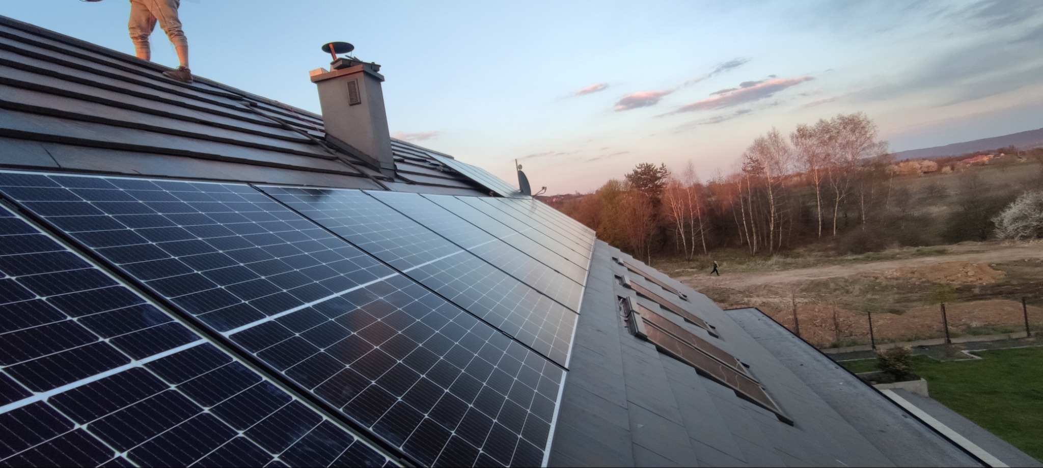 Instalacja paneli fotowoltaicznych na dachu domu jednorodzinnego z kominem, widok z góry, w tle zachodzące słońce i pejzaż.