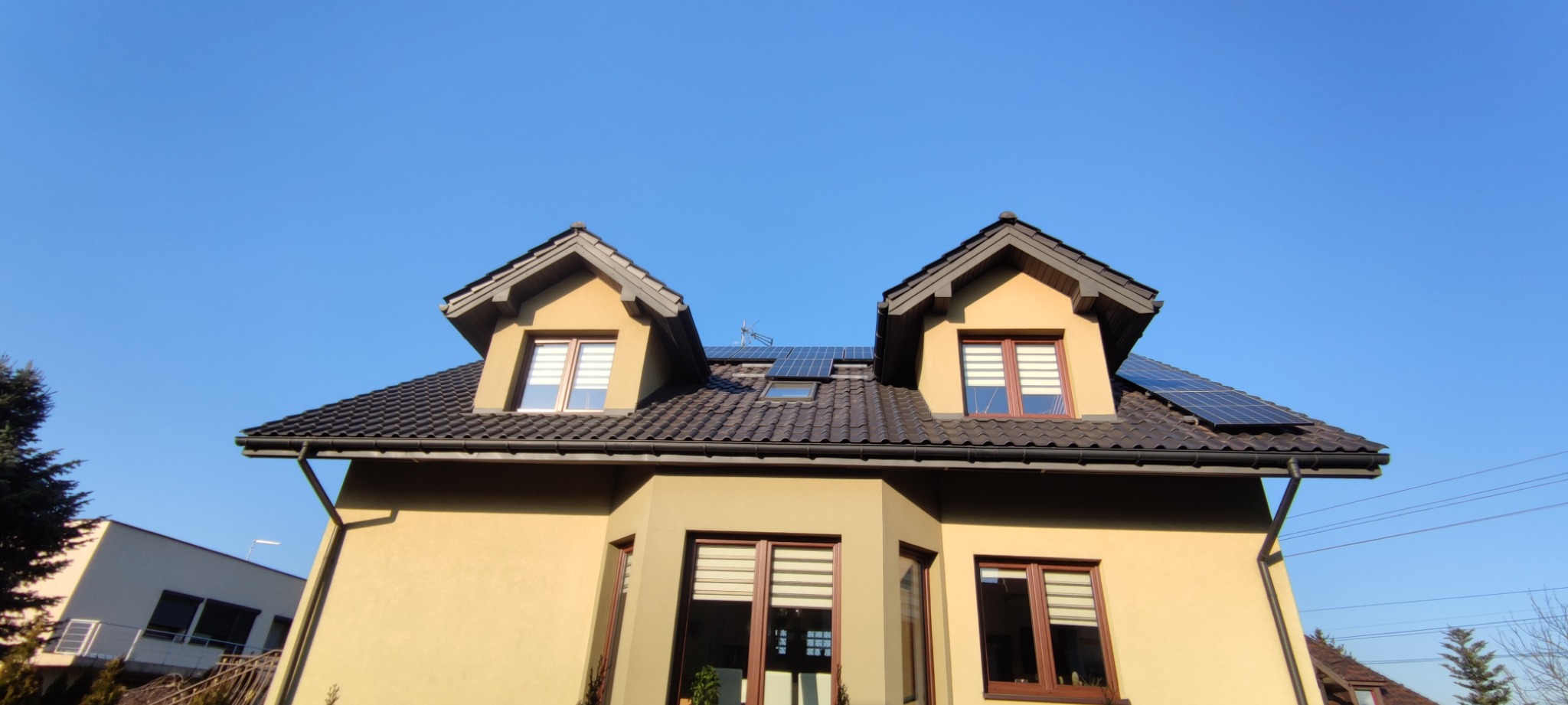 Dom jednorodzinny z brązowym dachem, oknami z roletami i panelami słonecznymi na dachu, uchwycony w słoneczny dzień pod kątem od dołu.