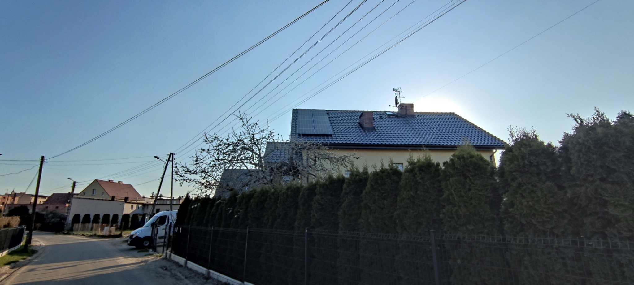 Dom jednorodzinny z panelami fotowoltaicznymi na dachu, widok z ulicy, zarośnięty żywopłot wzdłuż ogrodzenia, słoneczny dzień.