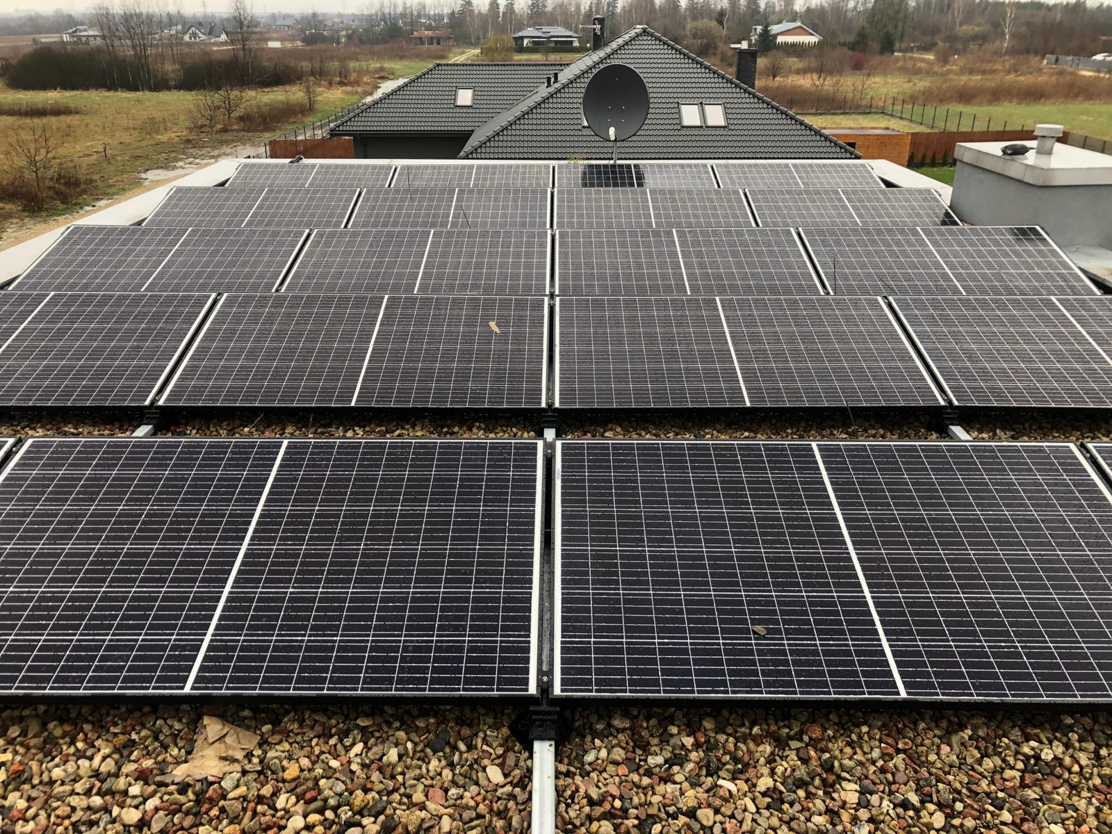 Widok z góry na panele fotowoltaiczne zamontowane na płaskim dachu pokrytym drobnym żwirem, z anteną satelitarną i budynkami w tle.