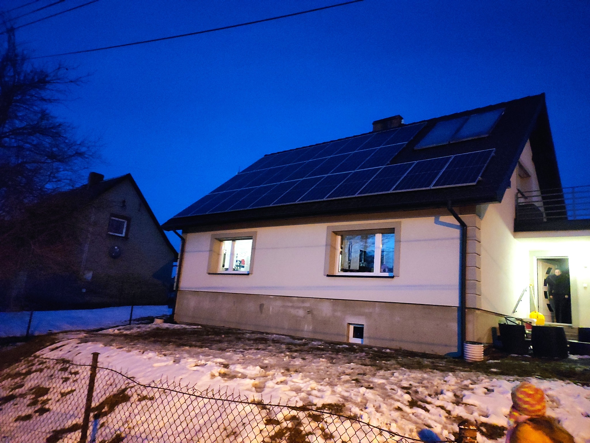 Dom jednorodzinny z panelami fotowoltaicznymi na dachu, widok z zewnątrz o zmierzchu, częściowo ośnieżony teren, osoba w drzwiach wejściowych.