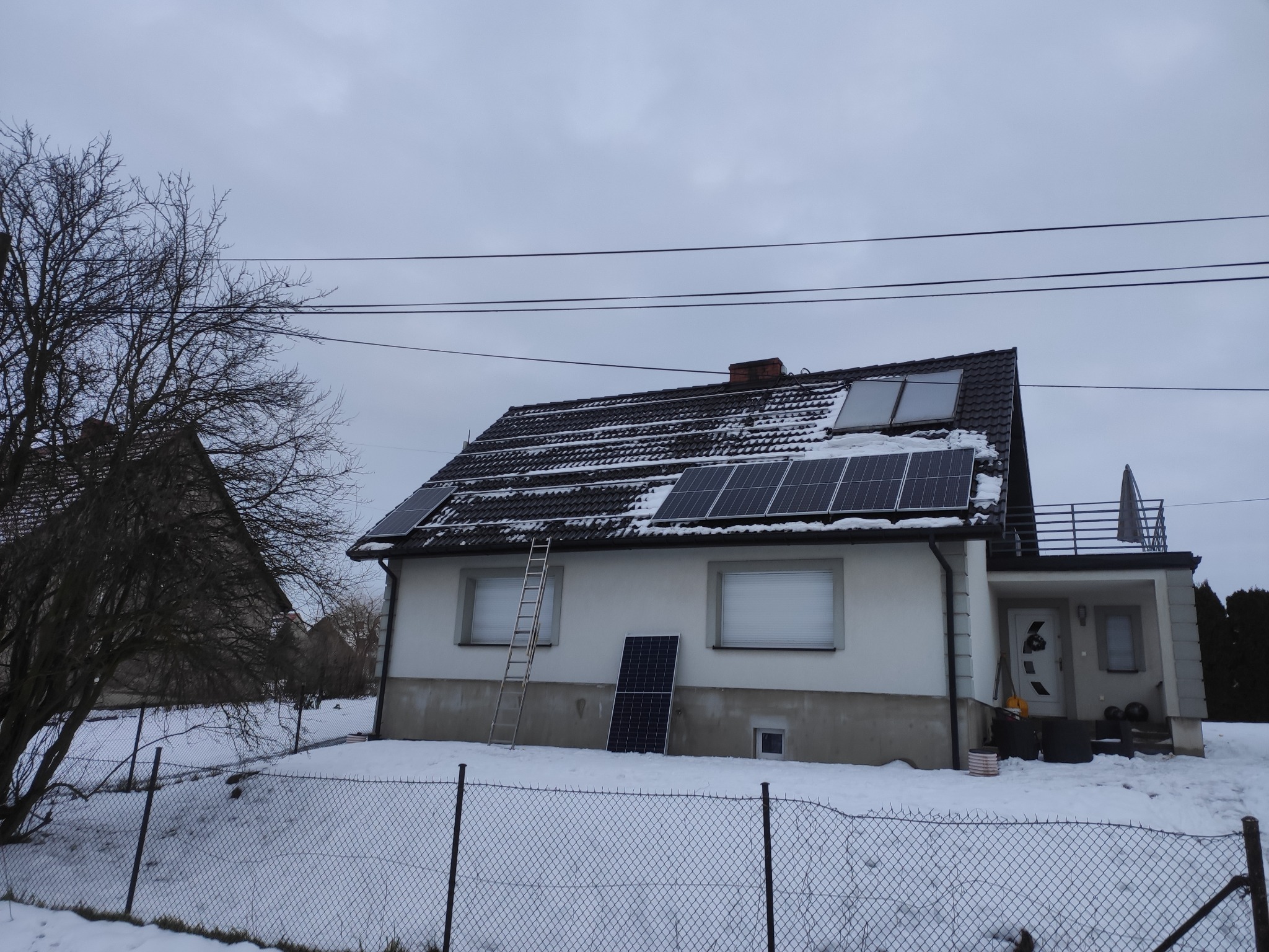 Dom jednorodzinny z panelami fotowoltaicznymi na dachu, drabina oparta o ścianę, pojedynczy panel oparty o ścianę, zimowy krajobraz.