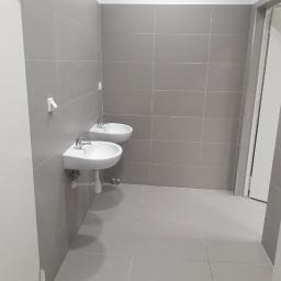 Firmus - Łazienka publiczna z dwoma umywalkami zamontowanymi na szarej, kafelkowanej ścianie i podłodze, widok z perspektywy wejścia.