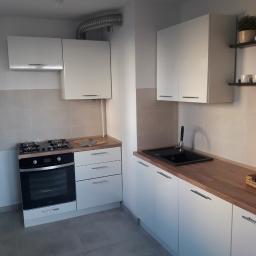 Firmus - Nowoczesna kuchnia z białymi szafkami, drewnianym blatem i czarnym zlewem, oświetlona lampą sufitową z trzema punktami świetlnymi.