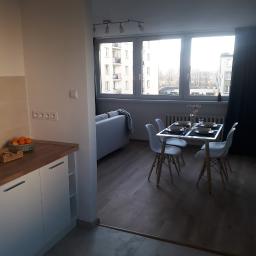 Firmus - Jasne wnętrze mieszkania z aneksem kuchennym, widok na stół z czterema krzesłami i okno z widokiem na budynek wielorodzinny.