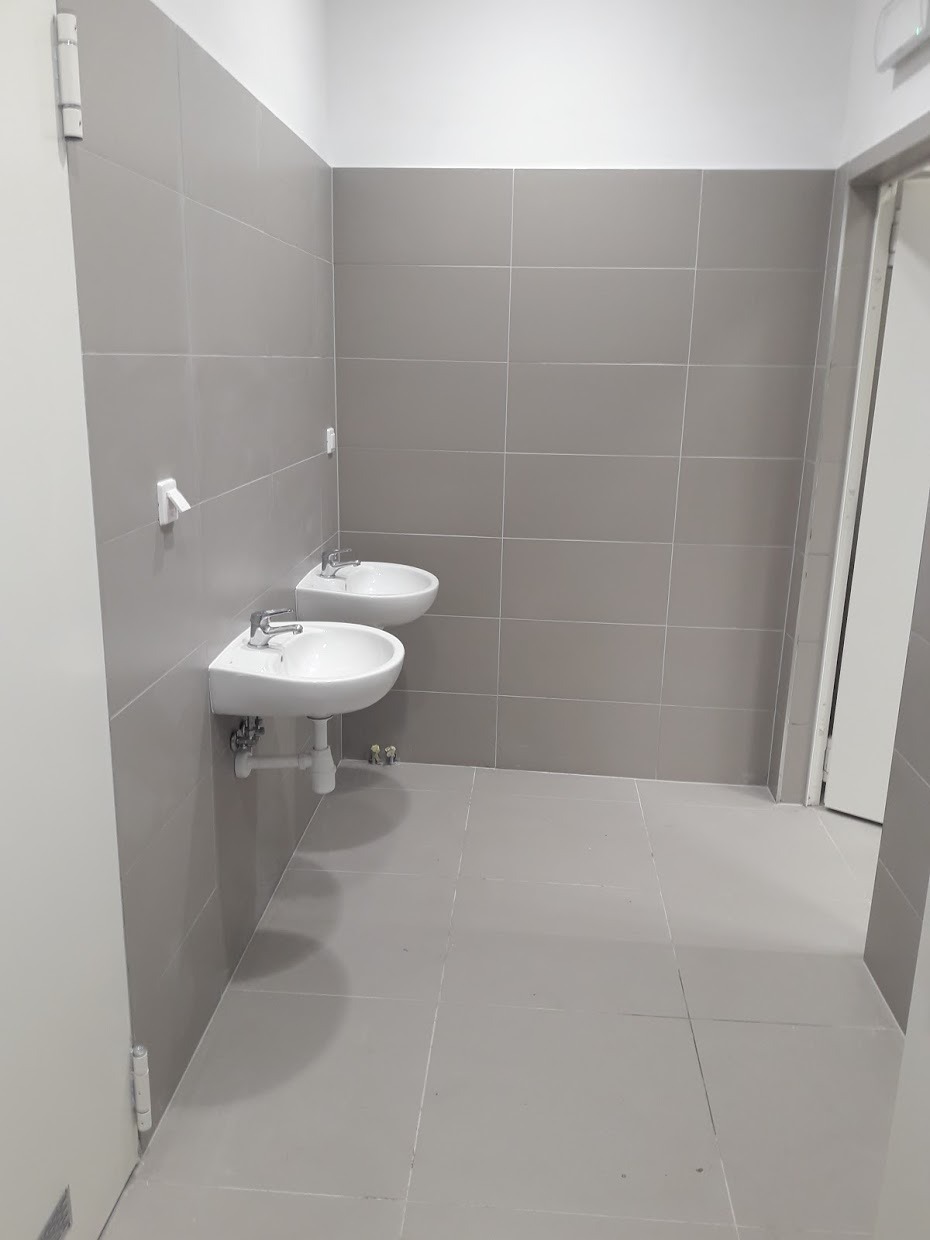 Łazienka publiczna z dwoma umywalkami zamontowanymi na szarej, kafelkowanej ścianie i podłodze, widok z perspektywy wejścia.