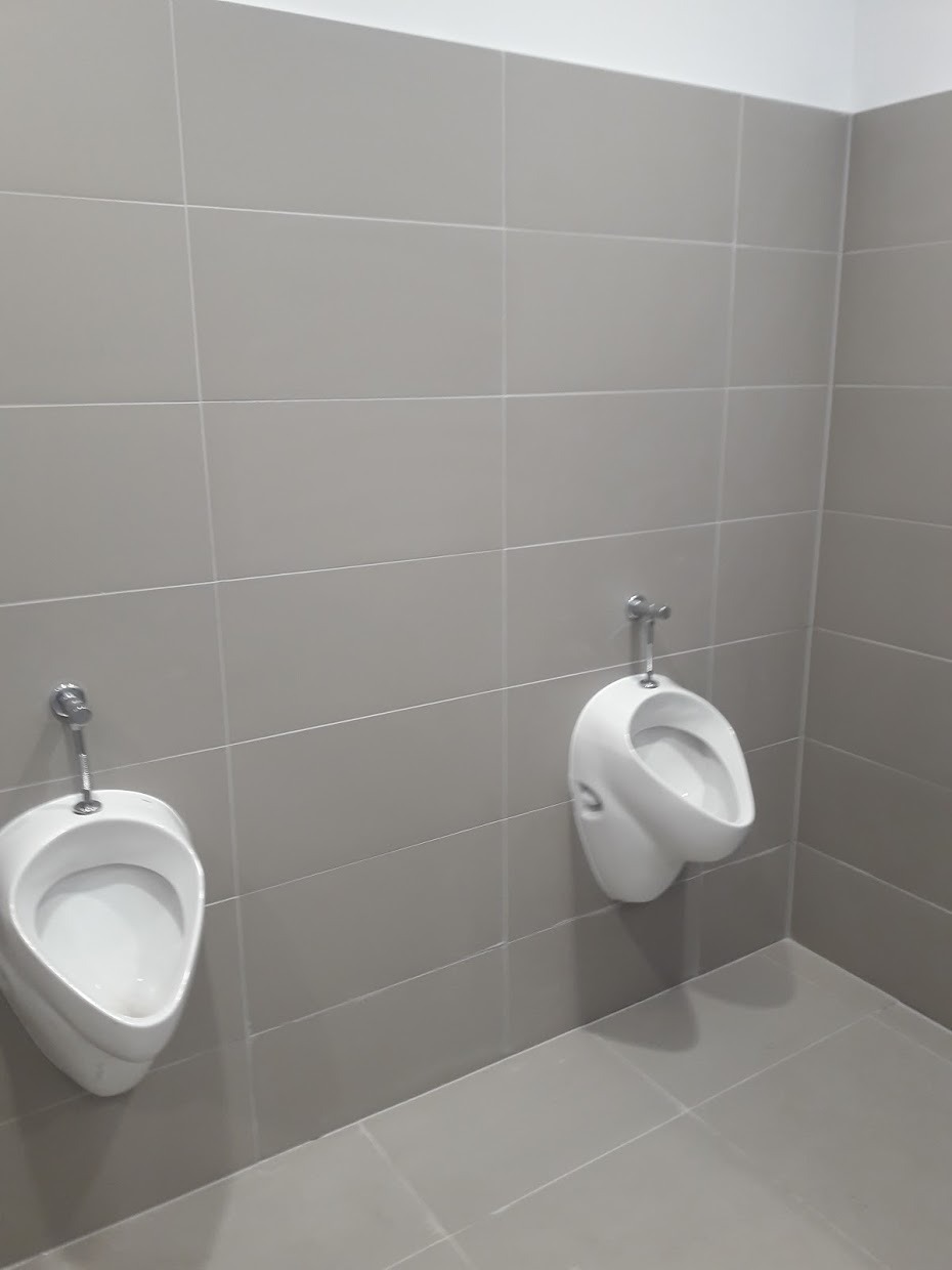 Dwa białe pisuary ceramiczne zamontowane na szarej, kwadratowej ścianie z płytek. Minimalistyczny wystrój toalety publicznej.