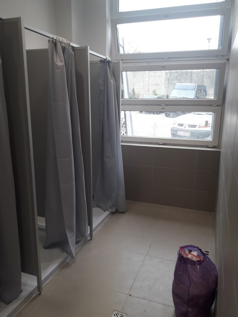 Publiczna łazienka z kabinami prysznicowymi oddzielonymi szarymi zasłonami, widok na zewnątrz przez okno, śmietnik z boku.