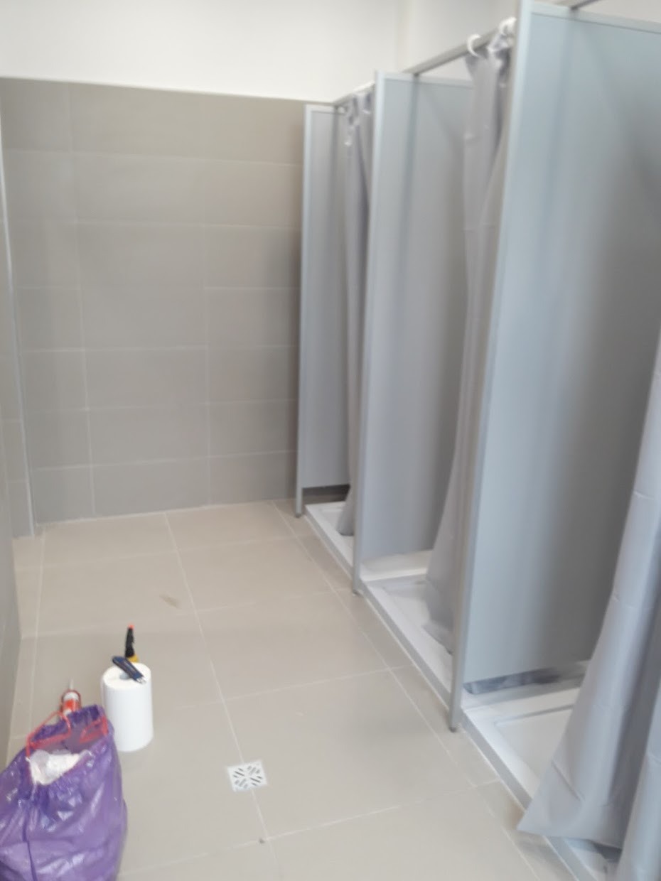 Szara łazienka publiczna z kabinami prysznicowymi oddzielonymi zasłonami, widoczne płytki na ścianach i podłodze, oraz torba z materiałami budowlanymi i narzędzia.
