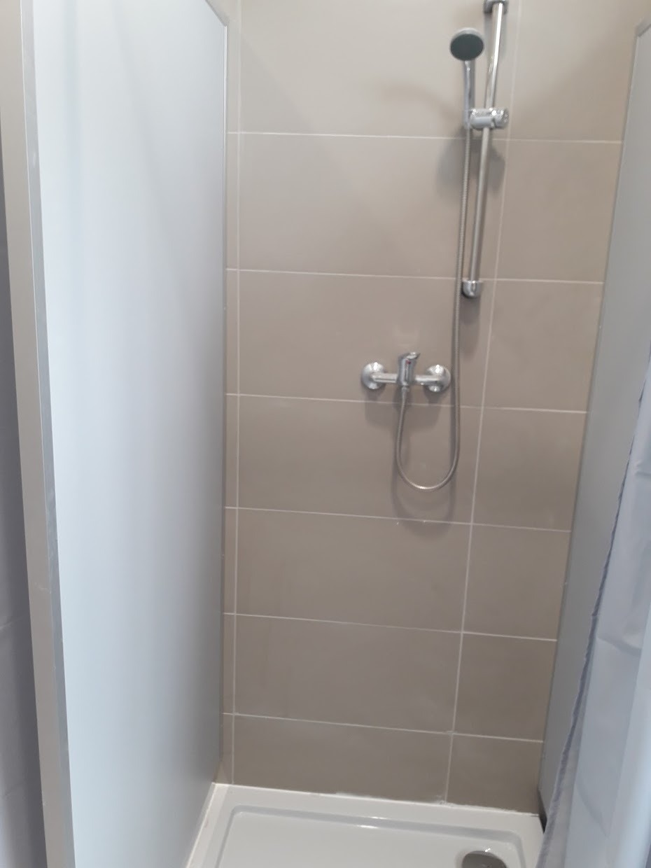 Beżowe płytki w kabinie prysznicowej z chromowaną armaturą i białą brodzikiem. Widoczny fragment białej ściany i zasłony prysznicowej.