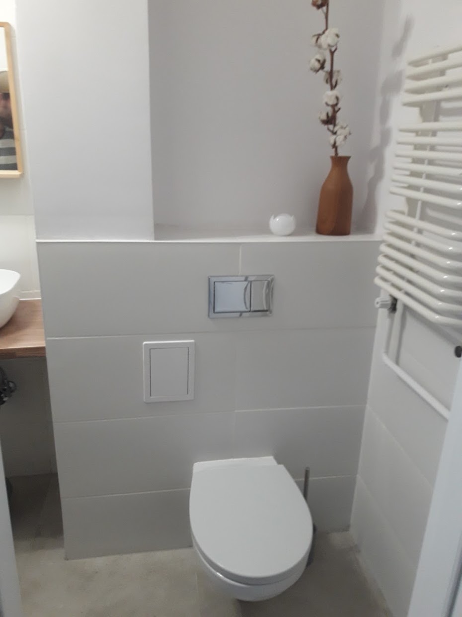 Nowoczesna toaleta podwieszana z białymi płytkami, ukrytym systemem spłukiwania i dekoracjami w postaci suszonych kwiatów w brązowym wazonie oraz białej kuli.