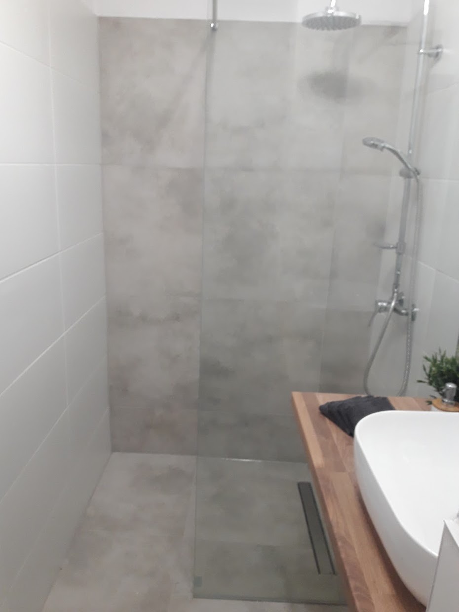 Nowoczesna kabina prysznicowa walk-in z szarymi płytkami imitującymi beton, drewnianym blatem pod umywalką nablatową i odpływem liniowym w podłodze.