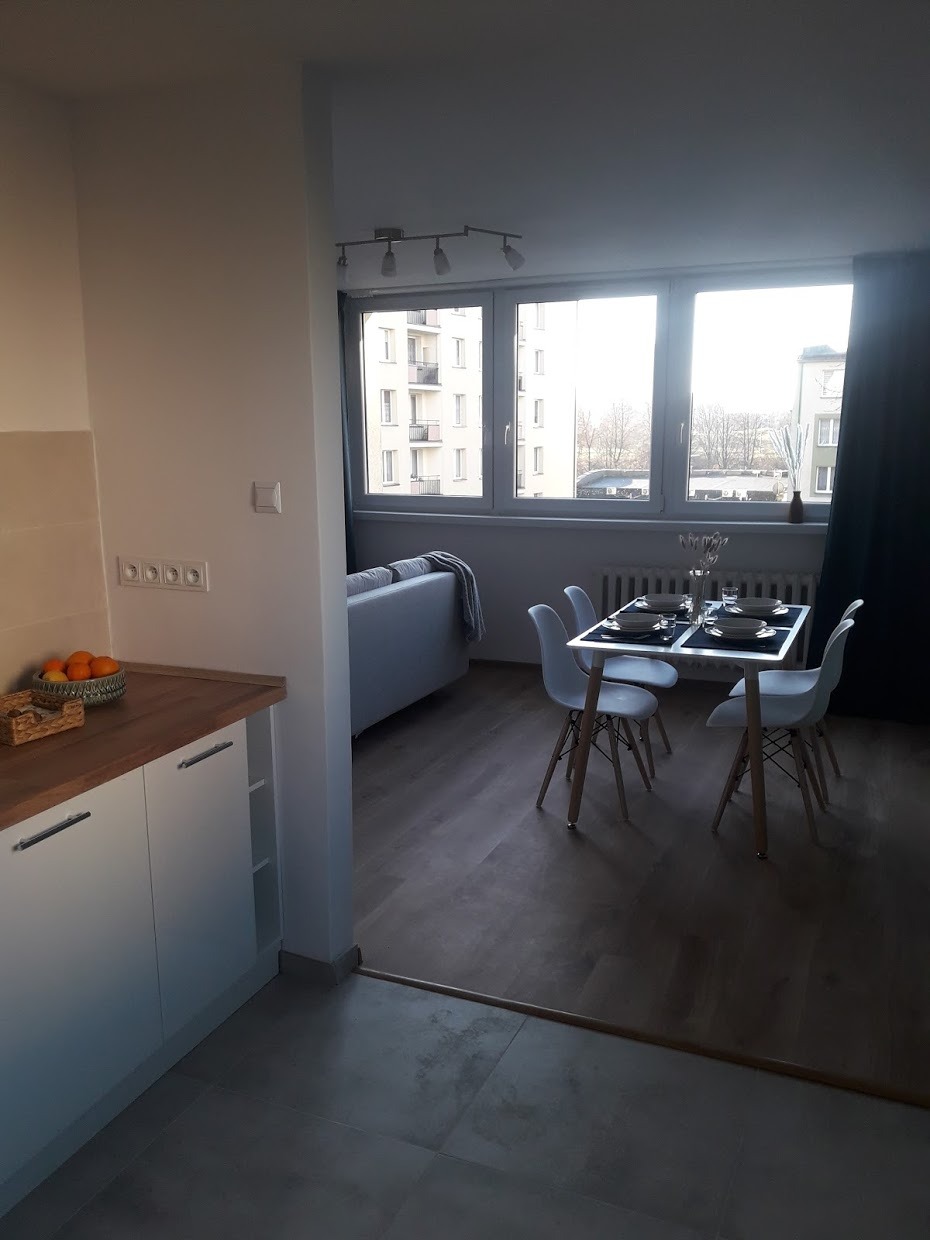 Jasne wnętrze mieszkania z aneksem kuchennym, widok na stół z czterema krzesłami i okno z widokiem na budynek wielorodzinny.