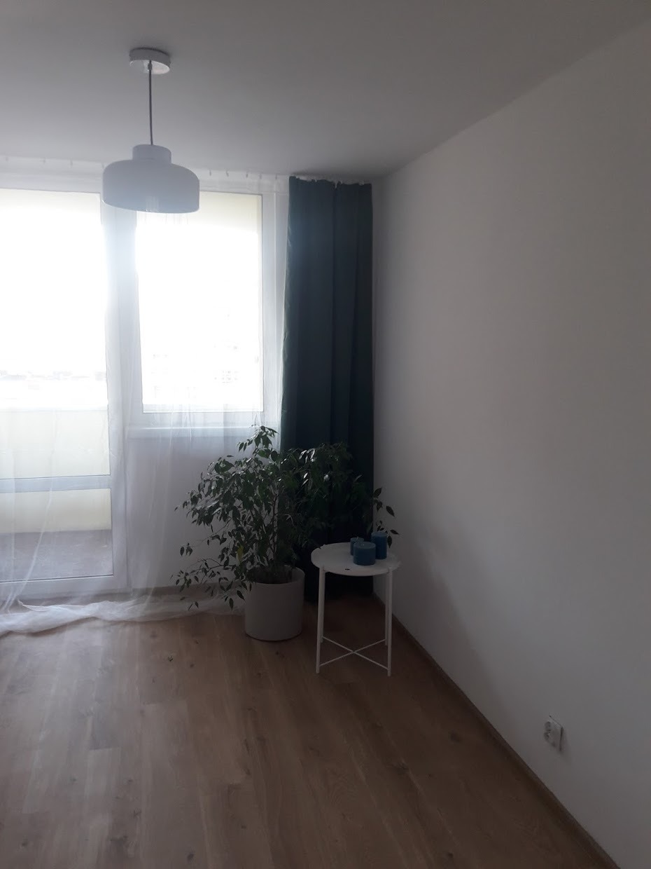 Jasne wnętrze po odświeżeniu: parkiet w odcieniach brązu, białe ściany, okno z firanką i ciemnozieloną zasłoną, roślina doniczkowa i mały stolik z dekoracjami.