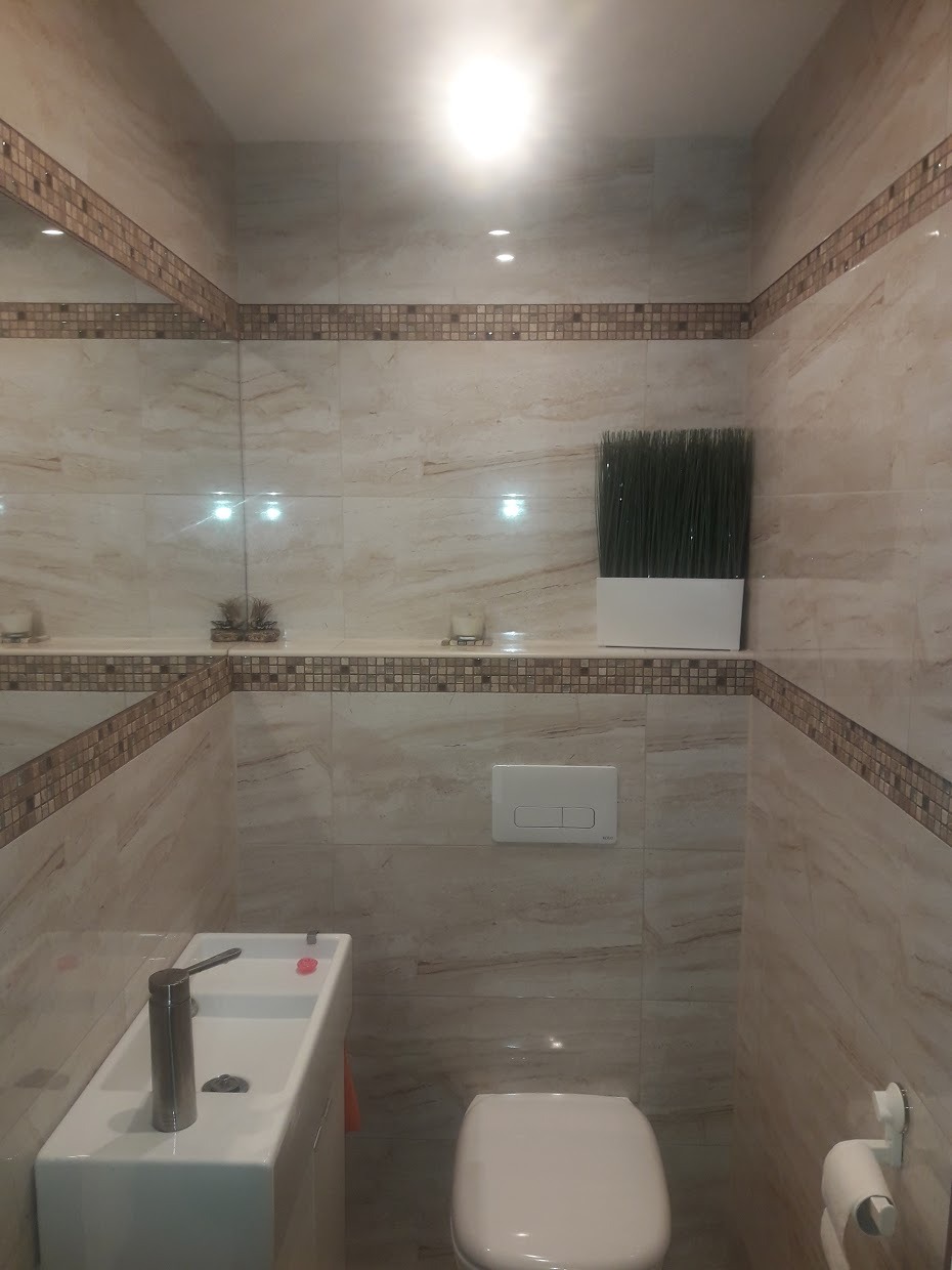 Jasna łazienka z beżowymi płytkami imitującymi marmur, dekoracyjnym pasem mozaiki, umywalką z szafką, toaletą i roślinną dekoracją na półce.