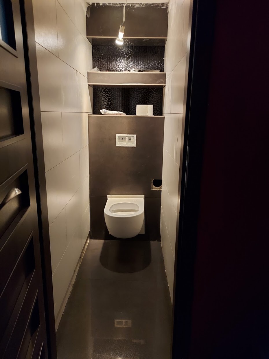 Wąska toaleta w trakcie remontu, widoczne płytki na ścianach, podwieszana miska WC i półki z czarną mozaiką, niedokończone prace wykończeniowe.