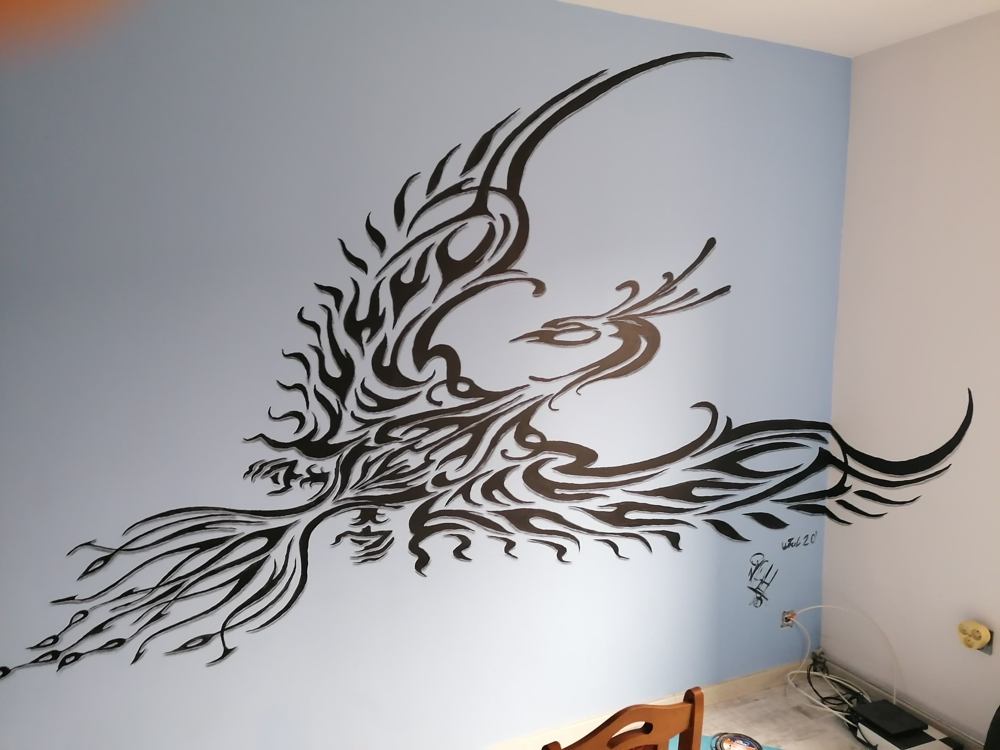 Czarny, ażurowy feniks na błękitnej ścianie, dekoracja w pokoju z widocznymi kablami i gniazdkami.