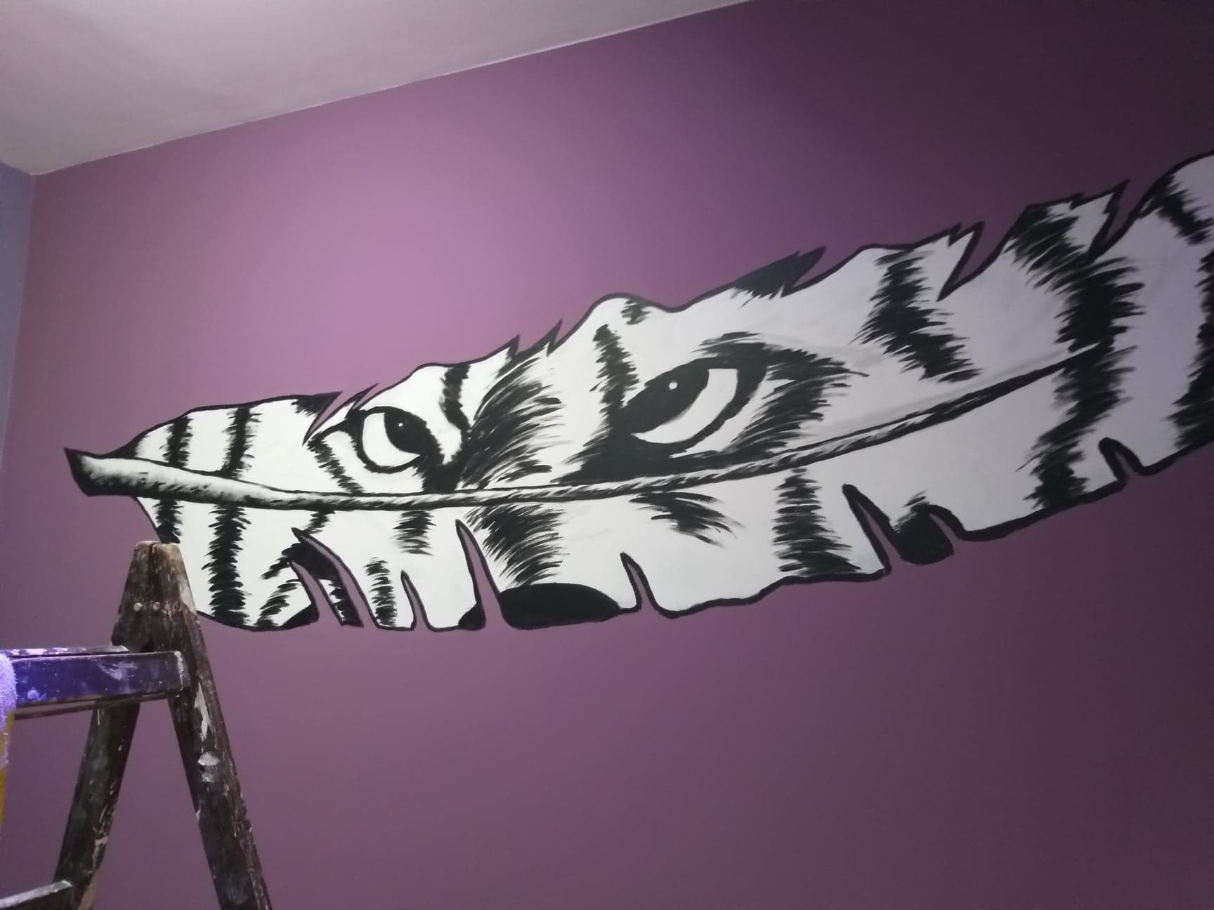 Ręcznie malowane na fioletowej ścianie pióro z motywem tygrysa, widoczna drabina malarska w lewym dolnym rogu.