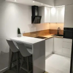 Nowoczesna, minimalistyczna kuchnia z białymi szafkami, drewnianym blatem i czarną armaturą, oświetleniem punktowym i wyspą z dwoma hokerami.