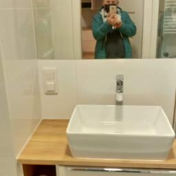 Mateusz Rokita - Biała, prostokątna umywalka nablatowa na drewnianym blacie, biała szafka pod umywalką, w lustrze odbicie osoby w masce robiącej zdjęcie telefonem.