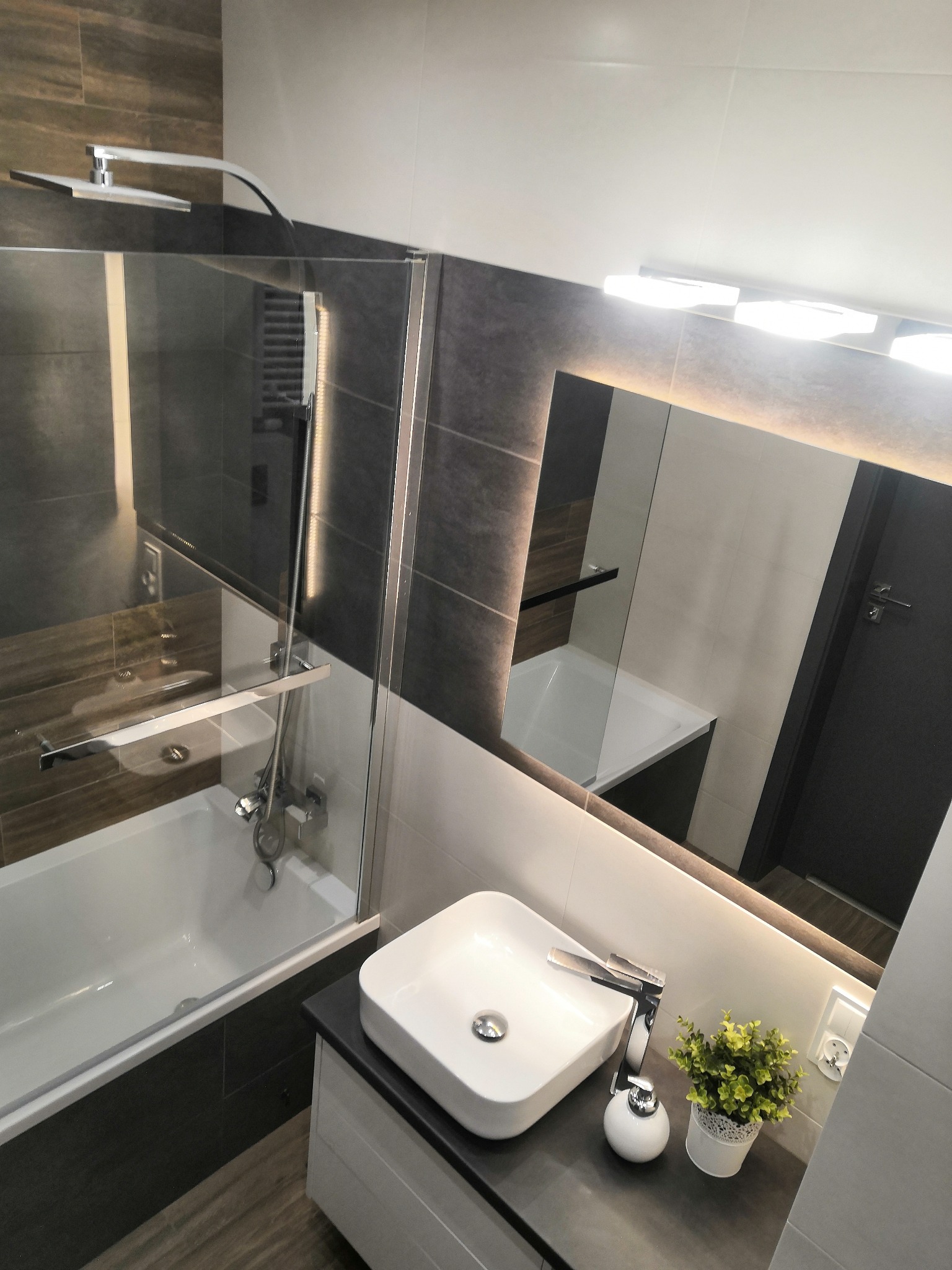 Nowoczesna łazienka z kwadratową umywalką nablatową, baterią, i dekoracyjną rośliną w doniczce na szarym blacie, widok na wannę za szklaną ścianką prysznicową i podświetlane lustro.