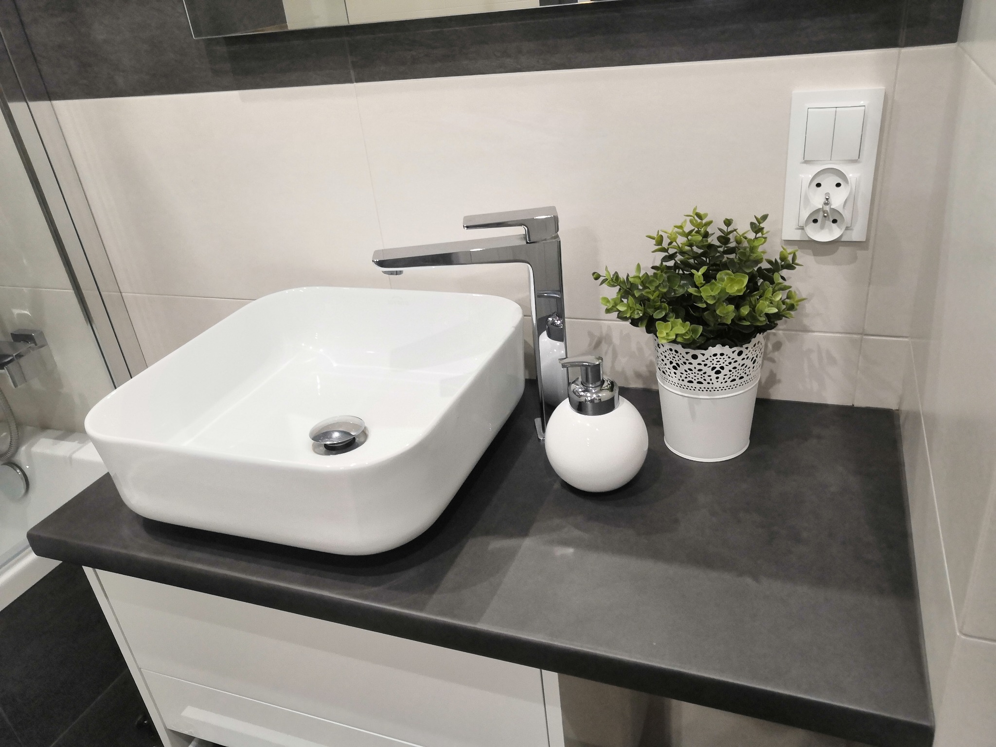 Biała, prostokątna umywalka nablatowa z chromowaną baterią, dozownikiem mydła i doniczką z zieloną rośliną na szarym blacie w nowoczesnej łazience z białymi płytkami i szarą dekoracyjną listwą.