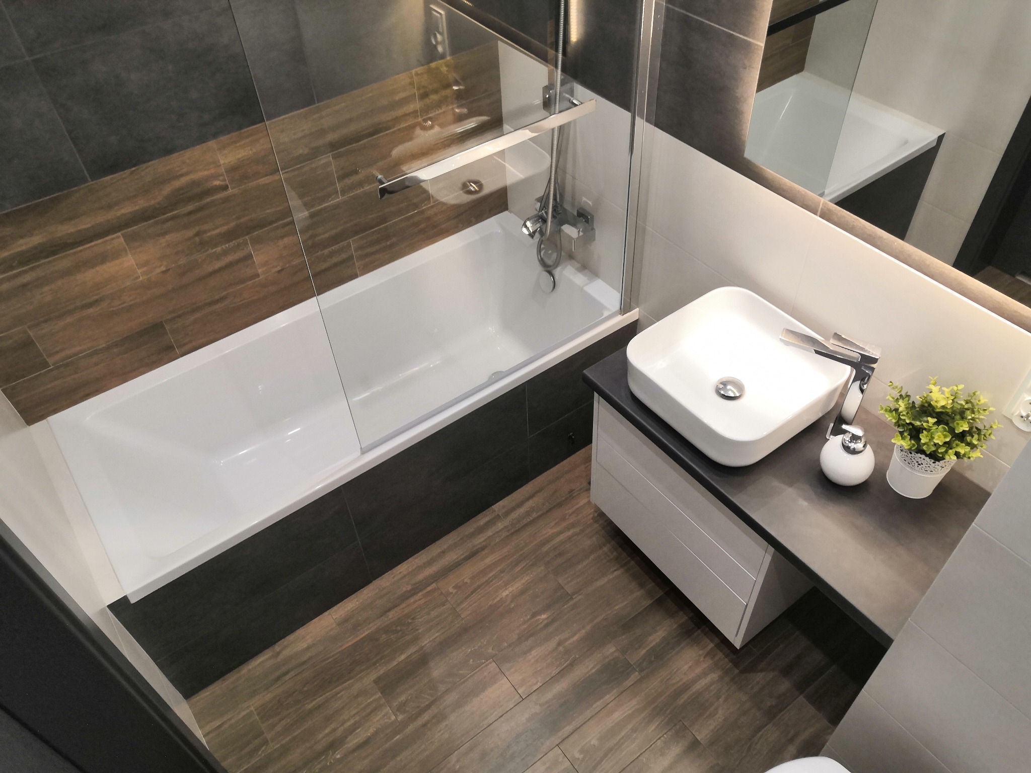 Nowoczesna łazienka z drewnopodobnymi płytkami, białą wanną za szklaną ścianką i umywalką nablatową z szafką w szarym odcieniu, widok z góry.