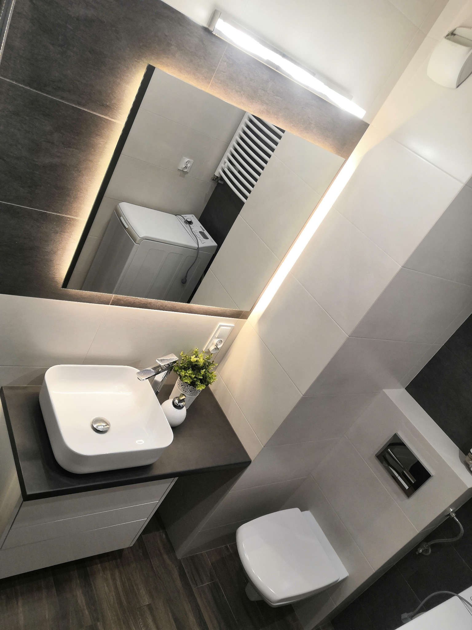 Nowoczesna łazienka z umywalką nablatową, podświetlanym lustrem, pralką widoczną w odbiciu i ciemną podłogą.