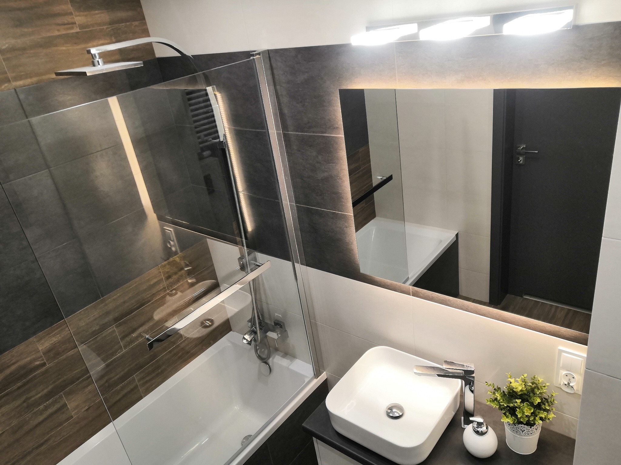 Nowoczesna łazienka z kabiną prysznicową typu walk-in, białą umywalką nablatową i lustrem z podświetleniem LED, wykończona płytkami w odcieniach szarości i brązu.