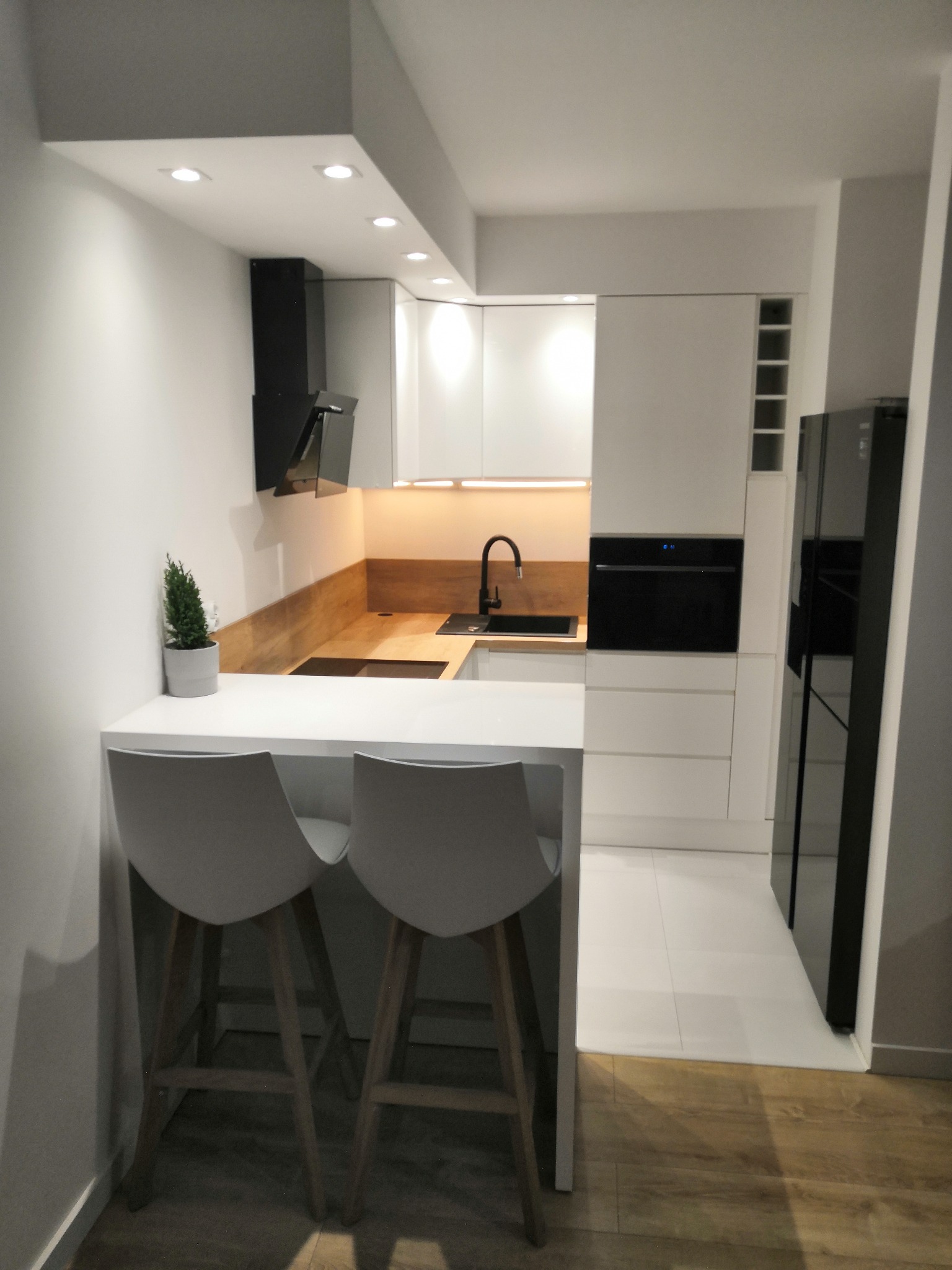 Nowoczesna, minimalistyczna kuchnia z białymi szafkami, drewnianym blatem, czarną armaturą i dwoma szarymi krzesłami barowymi z drewnianymi nogami, oświetlona punktowymi lampami