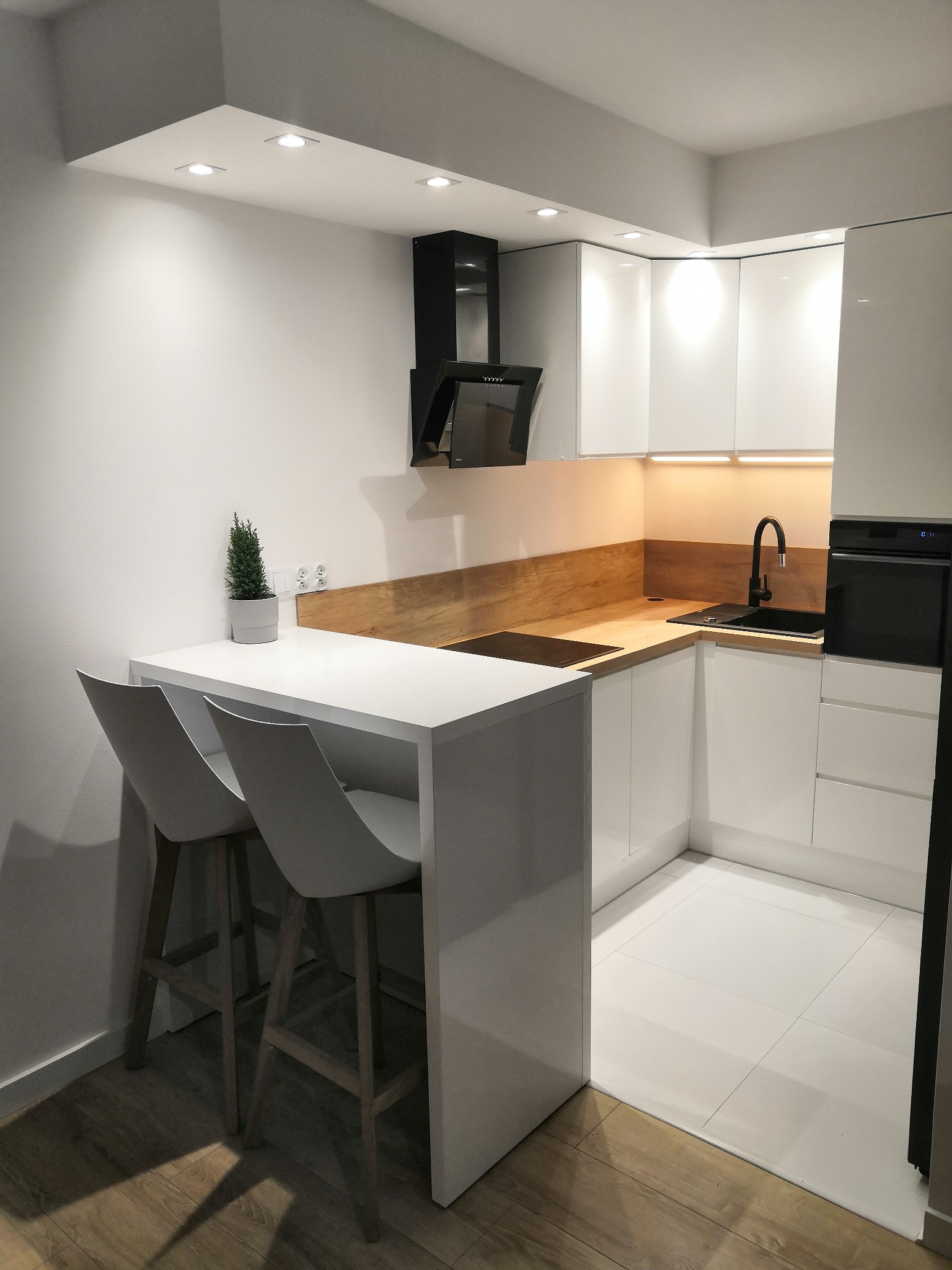 Nowoczesna, minimalistyczna kuchnia z białymi szafkami, drewnianym blatem i czarną armaturą, oświetleniem punktowym i wyspą z dwoma hokerami.