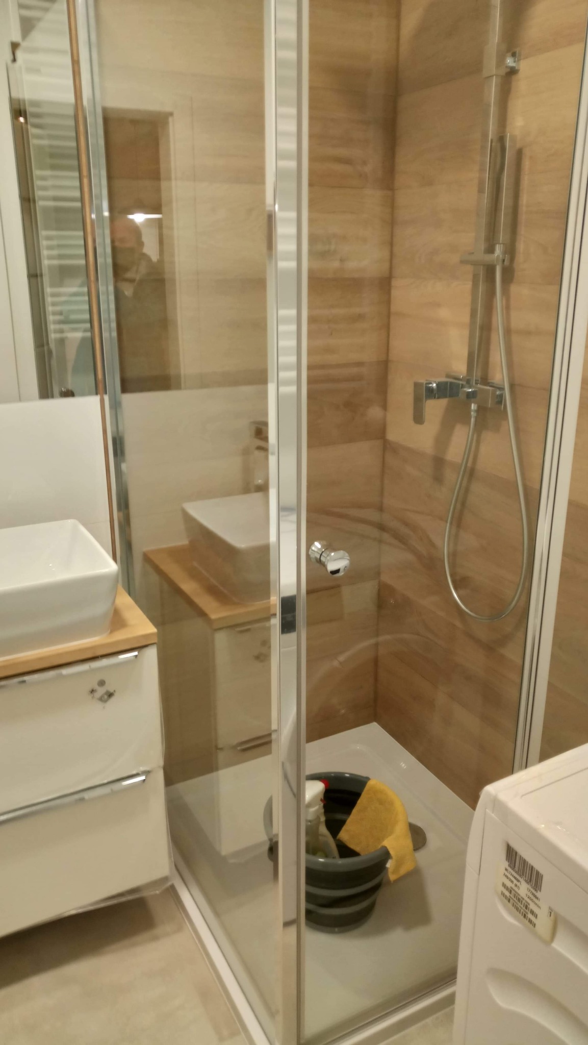 Nowoczesna kabina prysznicowa z drewnopodobnymi płytkami, widoczny fragment umywalki i szafki, wiadro z detergentami w brodziku, prawdopodobnie w trakcie sprzątania po remoncie.