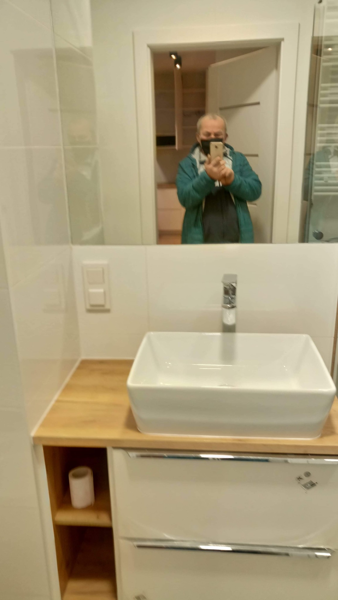 Biała, prostokątna umywalka nablatowa na drewnianym blacie, biała szafka pod umywalką, w lustrze odbicie osoby w masce robiącej zdjęcie telefonem.