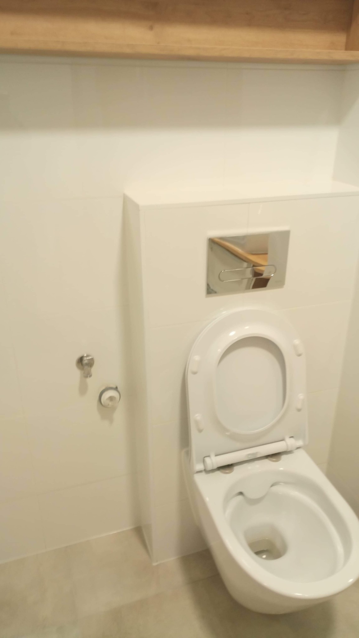 Biała, podwieszana toaleta z otwartą deską i chromowanym przyciskiem spłukiwania w minimalistycznej łazience wykończonej białymi płytkami i drewnianą półką.